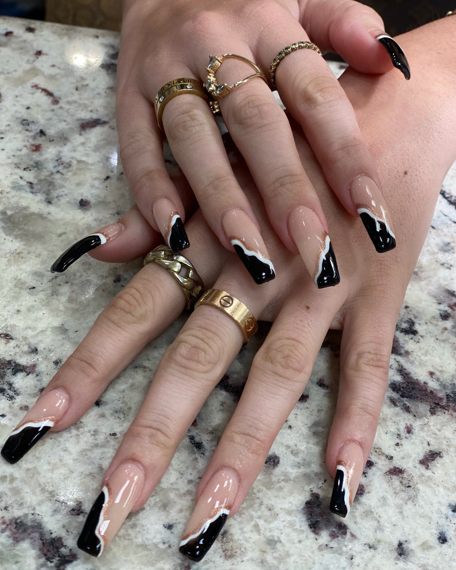A black stiletto nail set with white French detailing, worn with gold rings.