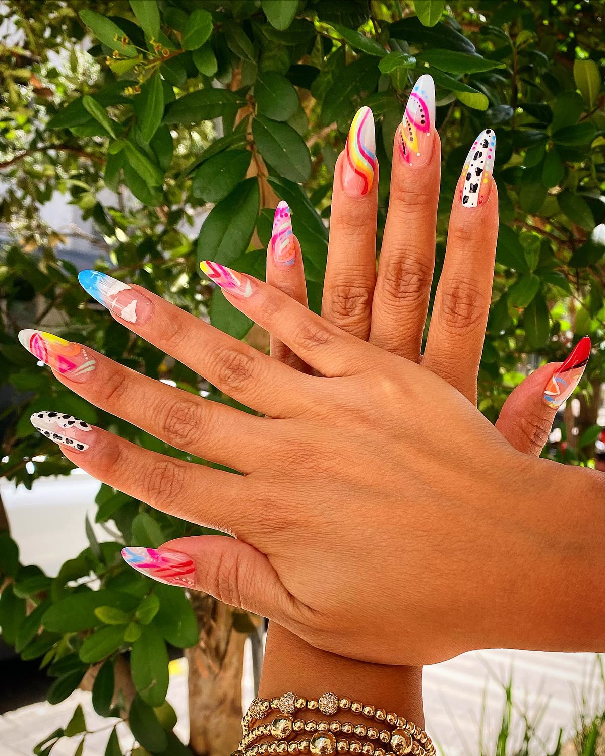 A hand with bright multicolour floral nail art held against garden greenery.