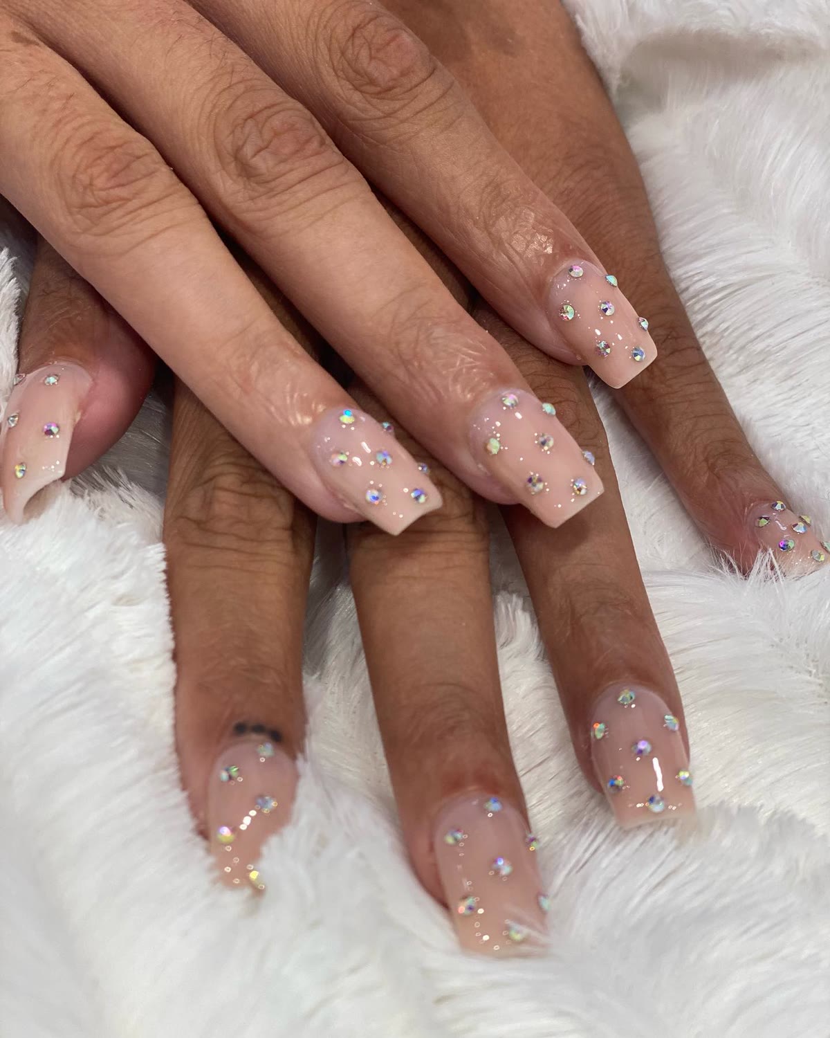 Long nude coffin nails with scattered rhinestone accents, resting on white fur.